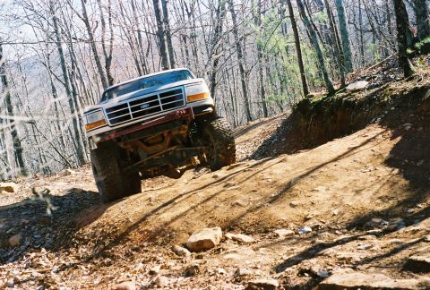 1978 Ford Bronco - White Ford F-Series truck navigates a steep, rocky trail through a wooded area.