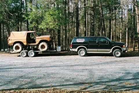 1978 Ford Bronco - A black SUV tows a vintage pickup truck on a trailer through a wooded area.