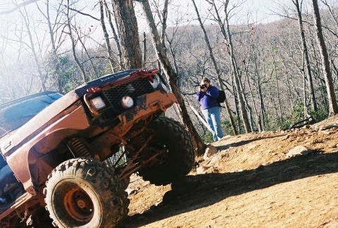 1978 Ford Bronco - A person photographs a heavily modified off-road vehicle navigating a challenging dirt path in a wooded area.