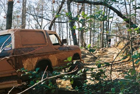 1978 Ford Bronco - A heavily muddied vehicle navigates a rough, wooded path with a person standing in the distance.