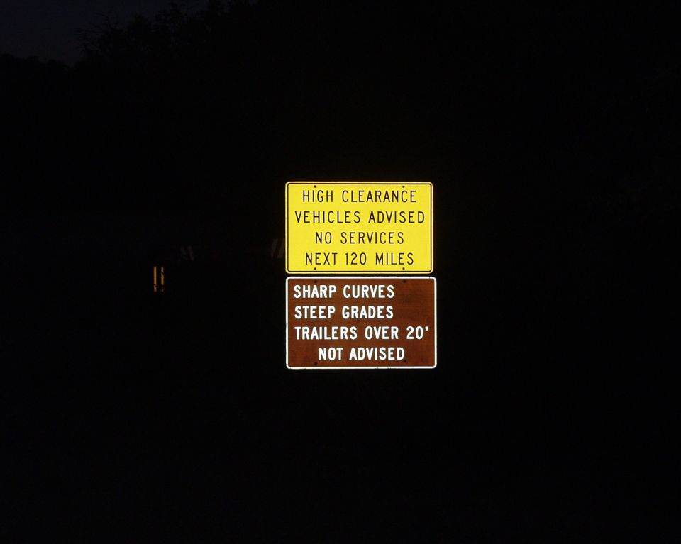Yellow and brown road signs warning of high clearance, no services, sharp curves, and steep grades