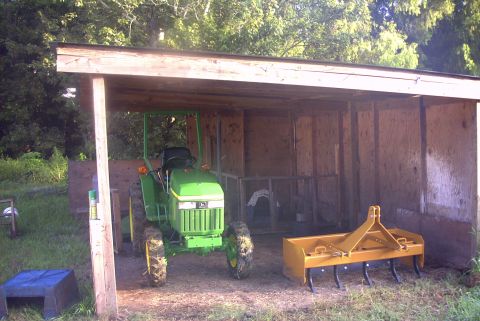 1984 Ford Bronco - A green tractor sits under a simple wooden shelter, with a yellow tiller attachment nearby.