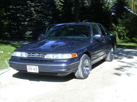 1996 Ford Crown Victoria - A dark blue sedan with chrome wheels is parked outside on a sunny day.