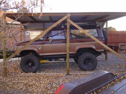 1986 Ford Bronco II - A brown Ford Bronco II is parked under a wooden shelter with a spare tire on the back.