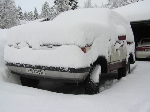 1989 Ford Bronco - A vehicle is heavily covered in snow, with only parts of the front and tires visible. Another car is parked in a garage in the background.