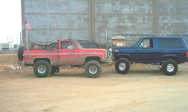 Dark blue lifted Ford Bronco parked beside a red topless lifted GM truck in front of a metal building