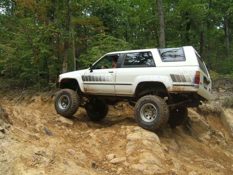 1993 Ford Bronco - A white off-road SUV tackles a challenging rocky incline on a wooded trail.