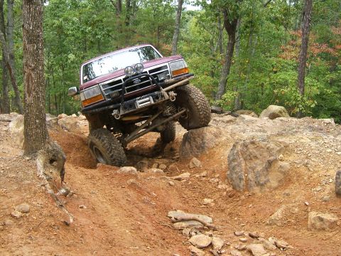 1993 Ford Bronco - A maroon Ford Bronco navigates a challenging rocky trail in a wooded area.