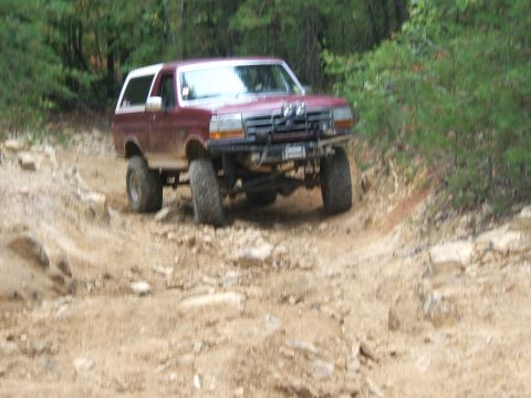 1993 Ford Bronco - A lifted Ford Bronco navigates a rocky, off-road trail surrounded by dense forest.