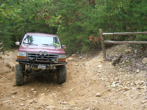 1993 Ford Bronco - A maroon Ford Bronco navigates a rocky, off-road trail surrounded by trees.