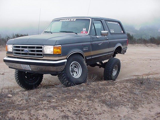 Gray lifted Ford Bronco on mud tires flexing on a dirt trail in foggy hills
