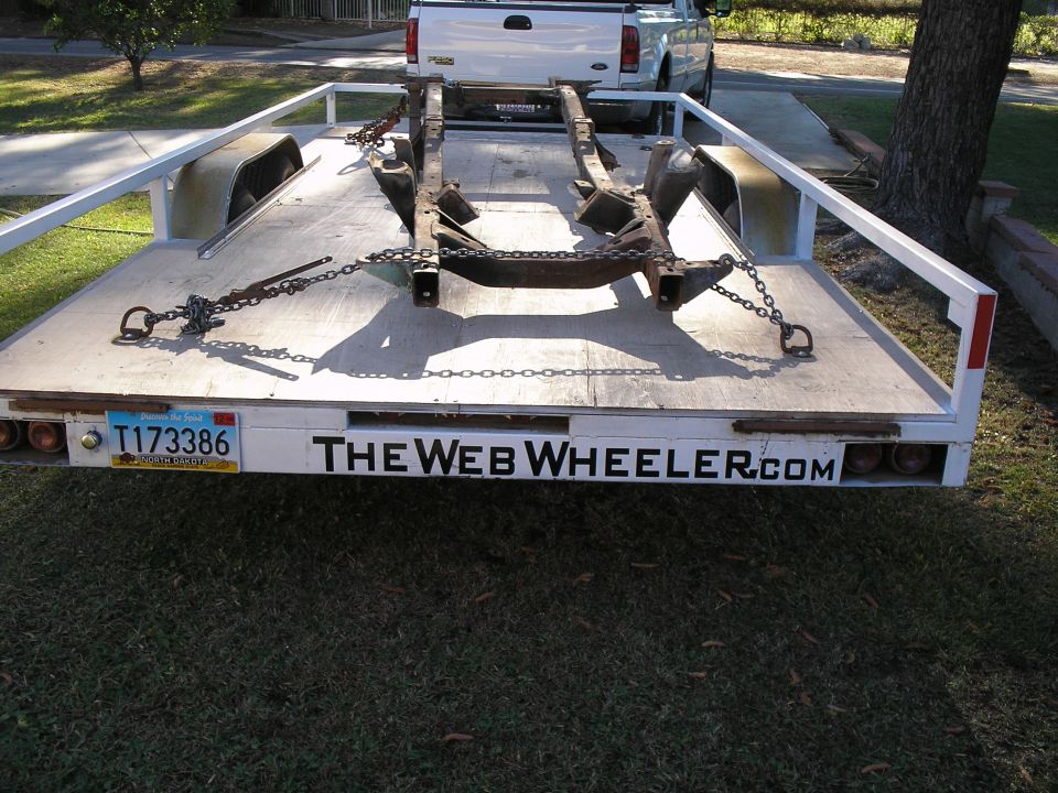 Trailer carrying a vehicle frame, with a white pickup truck behind it