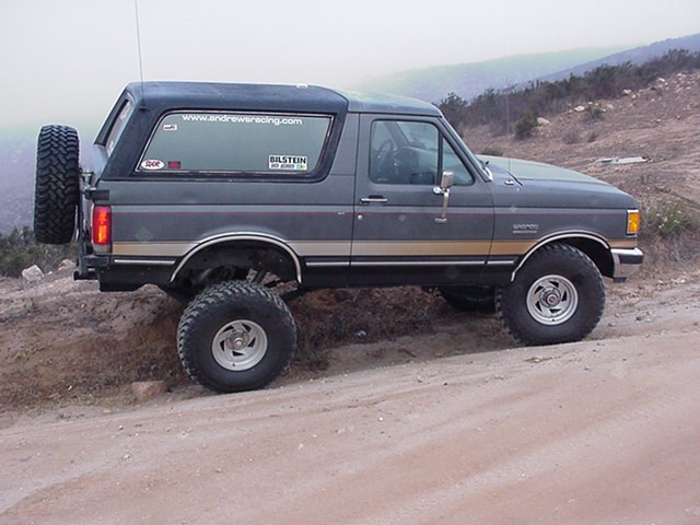 Side profile of gray lifted Ford Bronco with rear spare tire on a foggy dirt hillside