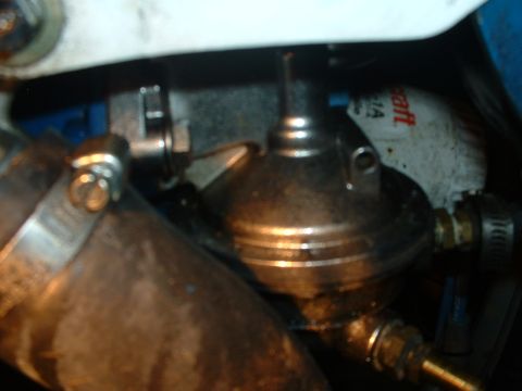 1985 Ford Bronco - Tight, slightly blurred engine-bay close-up of a bare-metal canister-style component with a vacuum nipple and brass fittings, mounted next…
