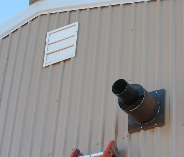 Rusty exhaust flue pipe through steel plate on tan metal shop wall beside white louvered vent