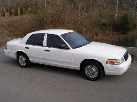 2001 Ford Crown Victoria - A white 2001 Ford Crown Victoria is parked on an asphalt road with trees in the background.