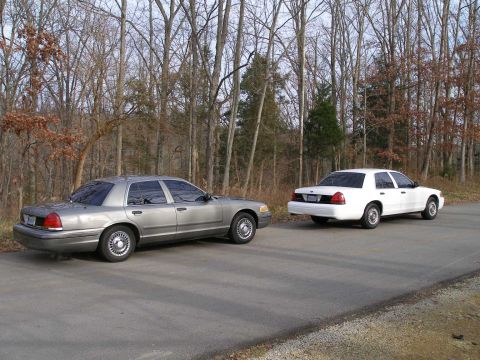 2001 Ford Crown Victoria - Two Ford Crown Victorias, a gray one and a white one, are parked side-by-side on a paved road with a wooded area behind them.