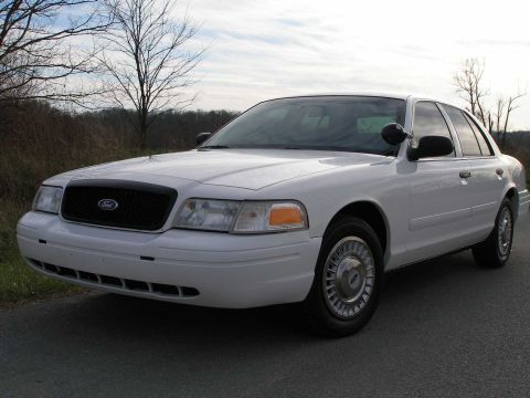 2001 Ford Crown Victoria - A white 2001 Ford Crown Victoria sedan is parked on a road with trees in the background.