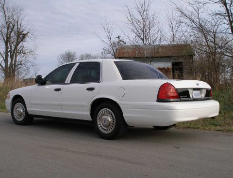 2001 Ford Crown Victoria - A white 2001 Ford Crown Victoria is parked on a road with a dilapidated building in the background.