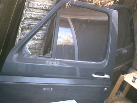 1990 Ford Bronco - Two dark grey/green truck doors stored on edge and leaning against a shed wall, glass and window frames removed from the nearer one, showin…
