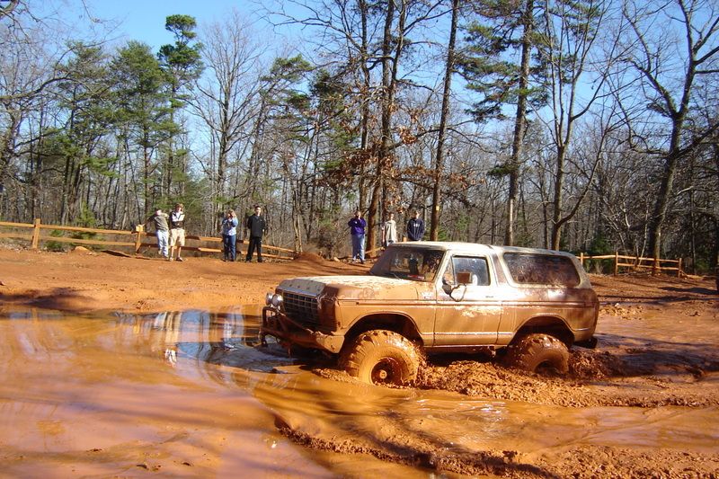 Brown Ford Bronco stuck in mud, front 3/4 view, off-road tires, trees and people in background