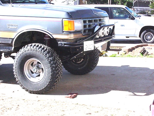 Front corner of gray Ford Bronco with black tube bumper, driving lights and skid plate