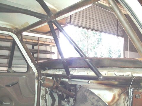 1988 Ford Bronco - Interior view of a stripped SUV cab showing a fabricated roll cage with black tubular front bars welded to bare metal roof structure. The d…