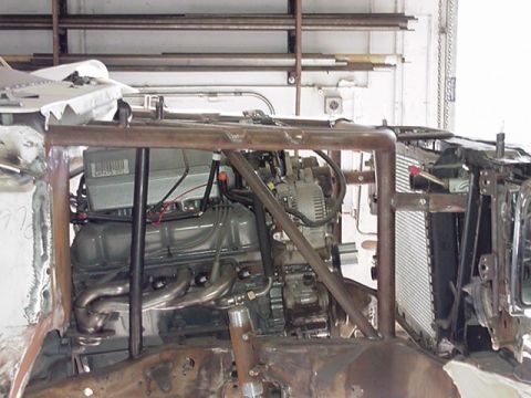 1988 Ford Bronco - Engine bay view of a vehicle mid-build, showing a V8 engine with exhaust headers, an alternator, and a custom tubular roll cage/frame struc…