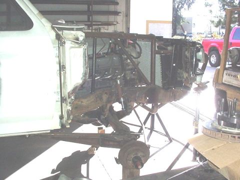 1988 Ford Bronco - A white Ford truck/SUV undergoing a major disassembly, shown from the front passenger side with the fender removed exposing the frame, fron…