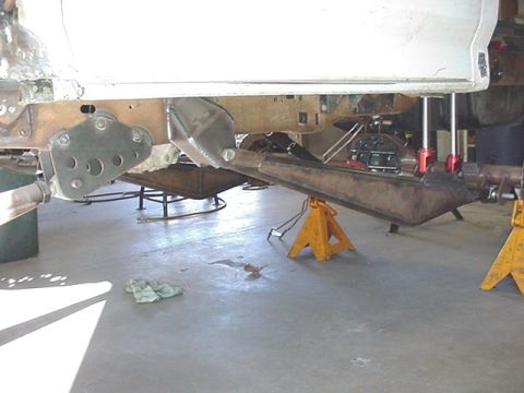 1988 Ford Bronco - Underside view of a vehicle raised on yellow jack stands, showing the rusty rear frame rails, a bare metal fuel tank or skid plate, and sus…