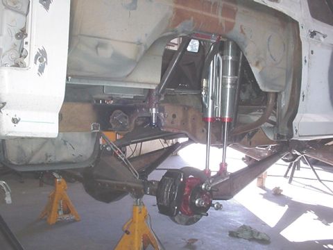1988 Ford Bronco - Underside view of a vehicle's rear suspension on jack stands, showing a fabricated four-link setup with dual polished coilover/shock units,…