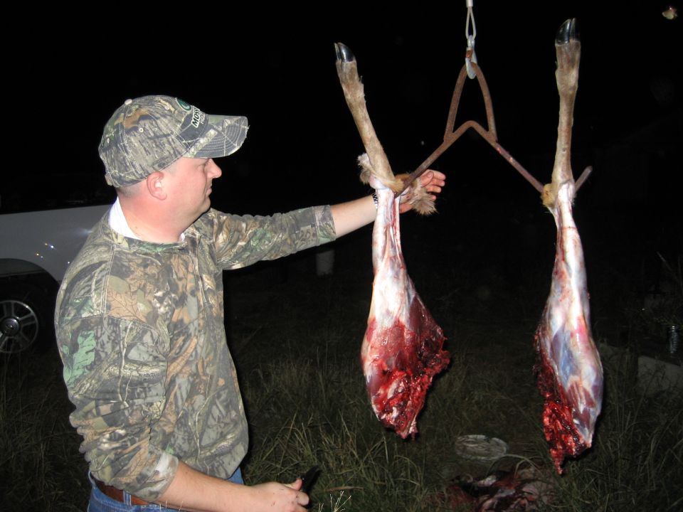 Man in camo holding two deer legs that have been skinned and are hanging from a metal spreader. A white truck is visible to the left