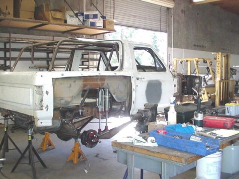 1988 Ford Bronco - A white Ford Bronco body shell undergoing restoration, viewed from the passenger side rear inside a workshop, sitting on jack stands with w…