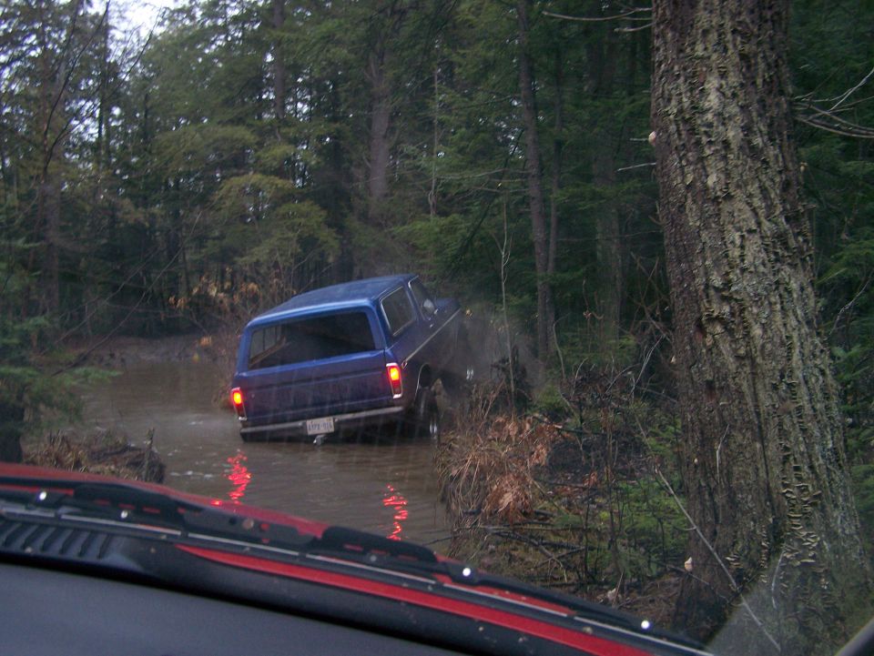 Blue SUV driving through muddy water in a forest. License plate is visible but unreadable
