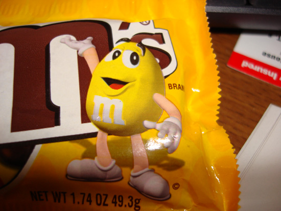 Yellow M&M character on a yellow M&M's bag, with text 'NET WT 1.74 OZ 49.3g' and 'SUPERMOTORS'