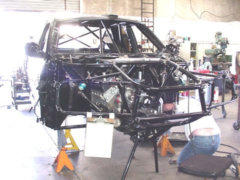 1988 Ford Bronco - A stripped vehicle body shell in a workshop, showing an elaborate black tubular roll cage and chassis framework with a V-engine mounted mid…