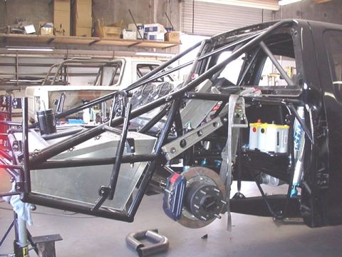 1988 Ford Bronco - Passenger-side rear view of a tube-chassis prerunner build in a shop, showing an extensive black roll cage, exposed front suspension with a…