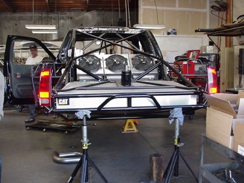 1988 Ford Bronco - Rear view of a tube-chassis prerunner-style truck build under construction in a workshop, showing a black roll cage, an aluminum bed with '…