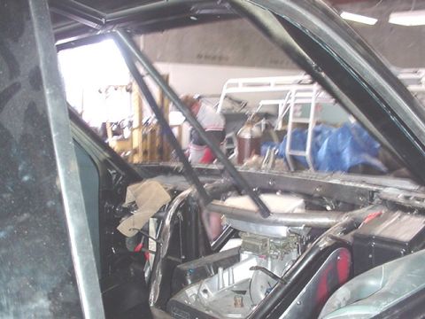 1988 Ford Bronco - Interior view through the side window opening of a vehicle with a bare metal roll cage, showing the tubular bars, an exposed engine area wi…