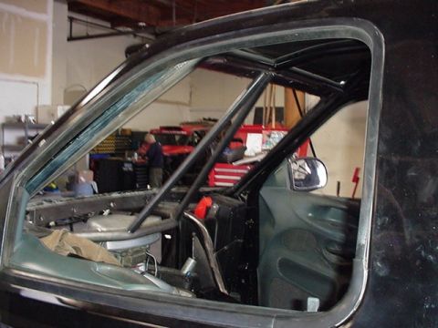 1988 Ford Bronco - View through the driver's side window opening of a dark-colored vehicle showing a welded tubular roll cage inside the stripped cab, with a…