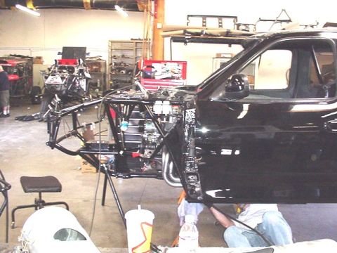 1988 Ford Bronco - A black pickup truck cab shell mounted alongside a bare tubular chassis undergoing a build inside a workshop, with an engine and components…