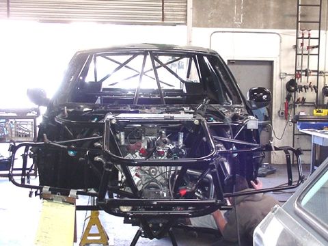 1988 Ford Bronco - Front view of a stripped SUV body shell mounted on a fabricated tubular chassis/frame with an exposed engine, inside a workshop with a pers…