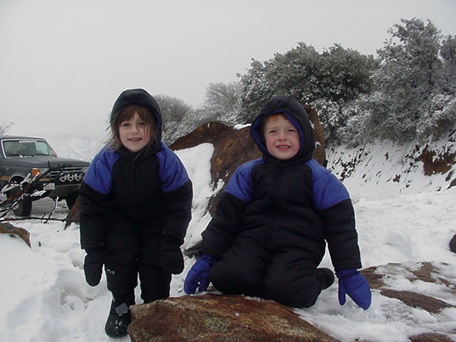 Two children in snowsuits posing on a rock in the snow