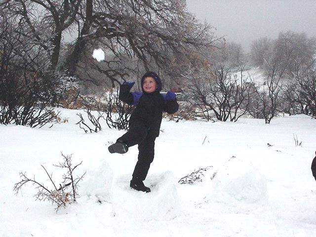 Child in a dark snowsuit throwing a snowball in a snow-covered field with bare trees
