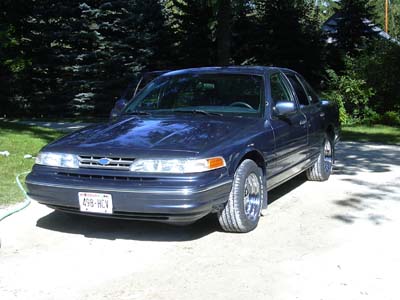 1996 Ford Crown Victoria - A dark blue sedan is parked on a gravel surface with trees in the background.