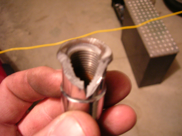 Close-up of a damaged lug nut with stripped threads, held by a hand. A yellow wire and a metal block are in the background