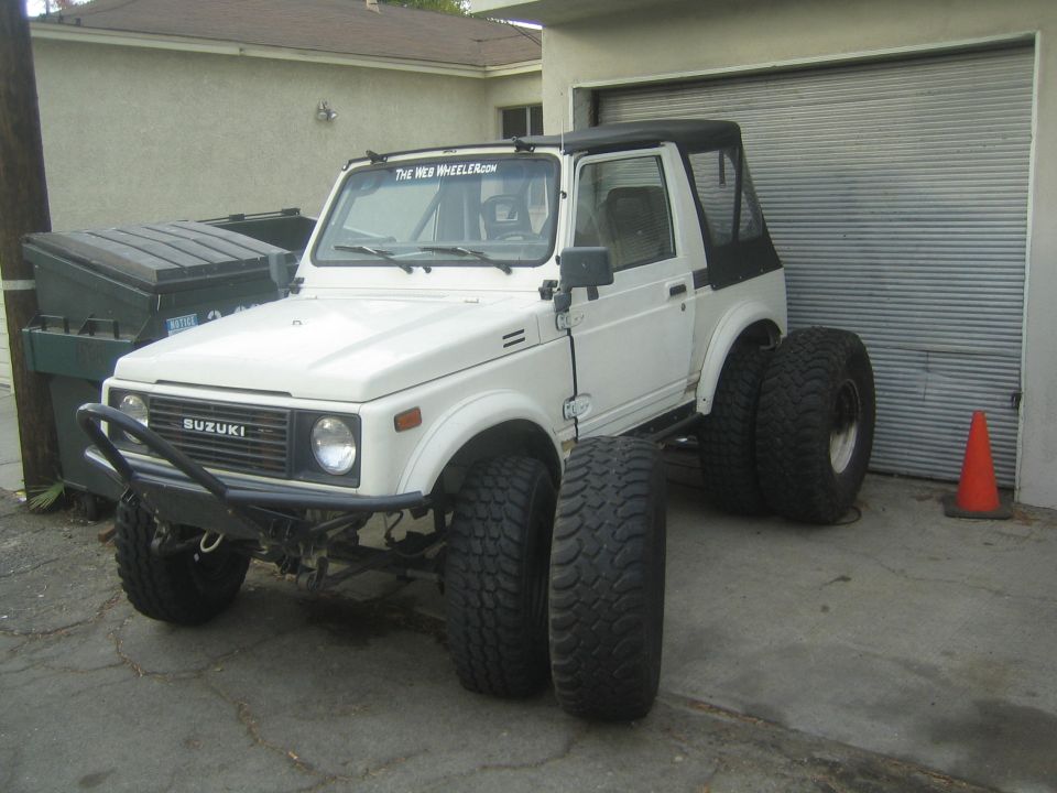 White Suzuki Samurai with black soft top and tube bumper, spare mud tires leaning against it by a garage