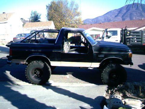 1989 Ford Bronco - This lifted Ford Bronco has a roll cage and is parked on a residential street with a white truck in the background.