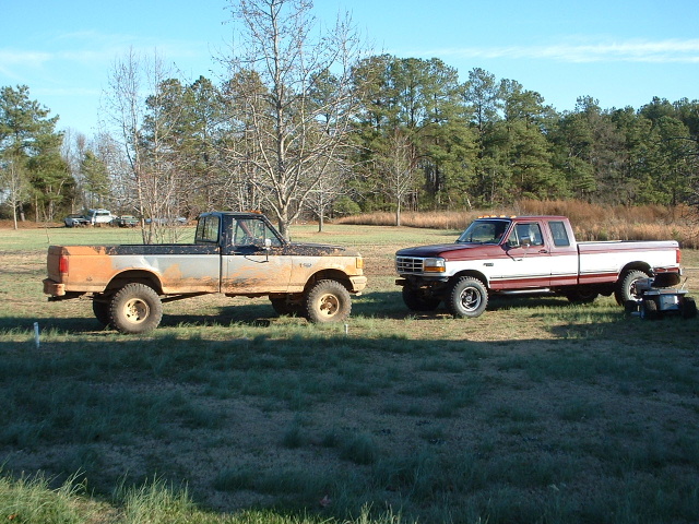 Two lifted Ford 4x4 pickups parked in a field, one orange primer and one red and white