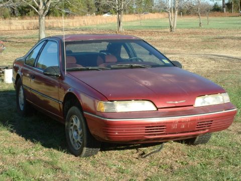 1989 Ford Thunderbird - A red 1989 Ford Thunderbird is parked outdoors on a grassy area with trees in the background.